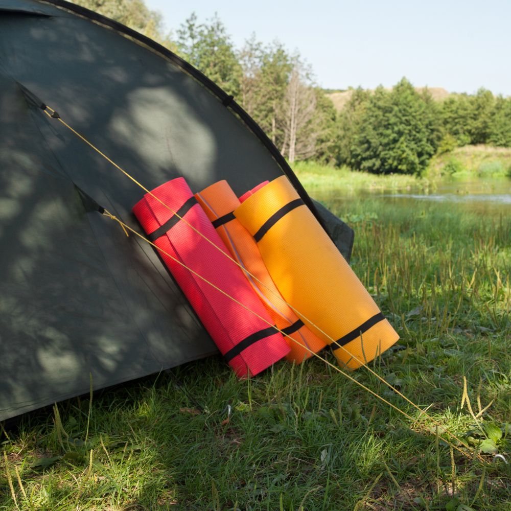 Camping scene with a tent and colorful sleeping pads on grass near a lake.