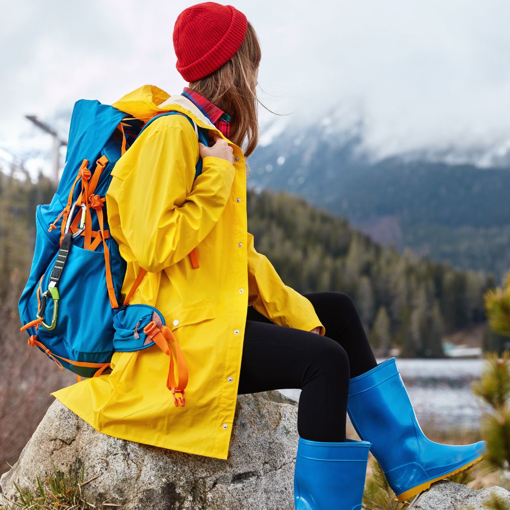 Person in yellow raincoat and blue boots with a red beanie and backpack sitting on a rock with mountains in the background.