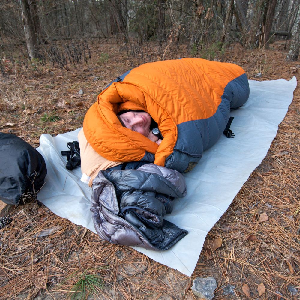 Person lying in an orange sleeping bag on a camping mat in a forest setting