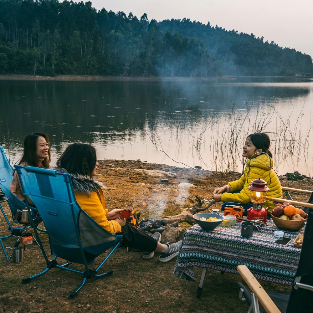 Three people sitting around a campfire by a lake, enjoying a picnic.